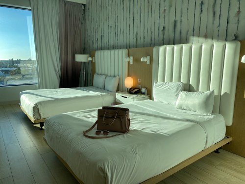 A modern hotel room with two beds, a window with natural light, and a bag and sunglasses on the bed.