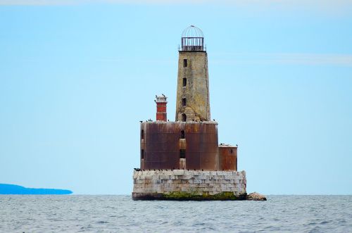 A weathered lighthouse stands on a rocky island, surrounded by calm waters under a clear blue sky.
