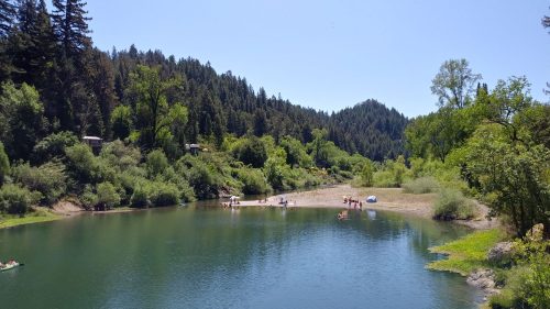 A serene river scene surrounded by lush green trees, with people enjoying the water and sandy beach under a clear blue sky.