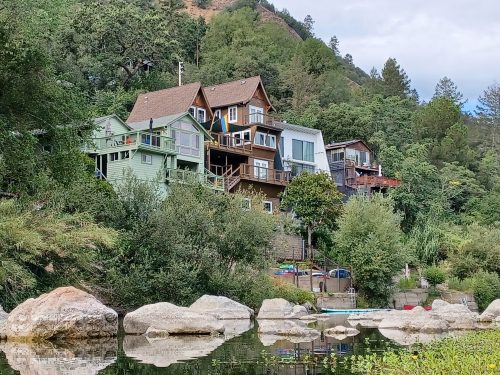 A hillside with colorful houses overlooking a calm river, surrounded by lush greenery and large rocks.