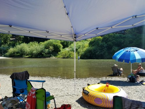A shaded area by a river with chairs, an inflatable tube, and people relaxing under umbrellas. Green trees in the background.