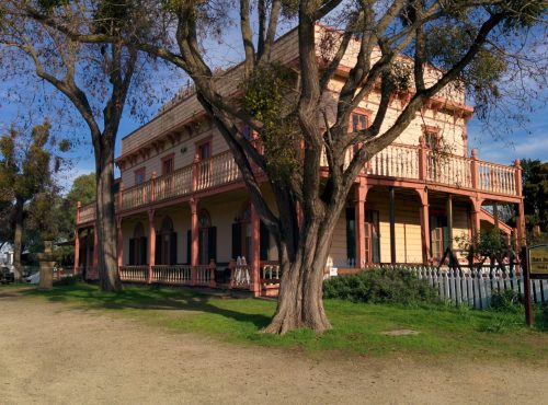 A historic two-story building with a porch, surrounded by trees and greenery, set in a rural landscape.