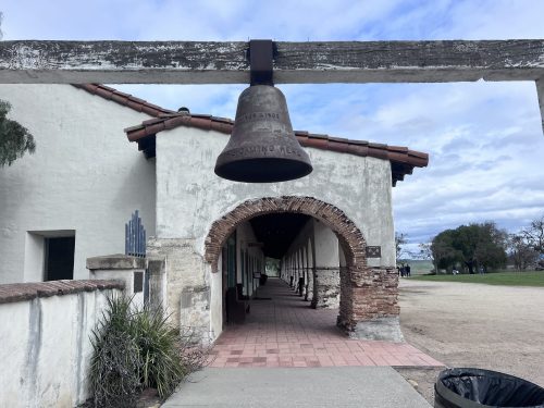 A large bell hangs from a wooden beam above an archway leading to a historic building with a tiled roof.