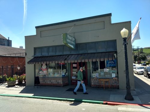 A quaint grocery store with a bakery sign, featuring a colorful storefront and a person walking by.