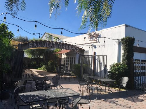 Entrance to San Juan Gardens with string lights, surrounded by greenery and outdoor seating. Clear blue sky above.