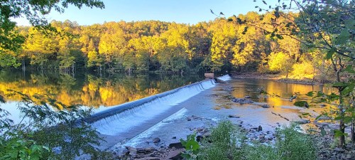 A serene river scene with a small dam, surrounded by vibrant autumn foliage and calm water reflecting the trees.