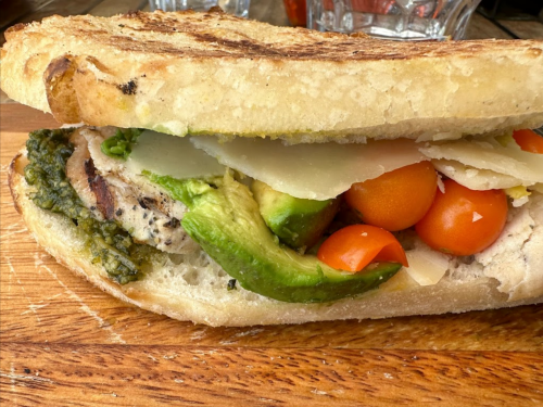A close-up of a sandwich with grilled chicken, avocado, cherry tomatoes, and pesto on toasted bread.