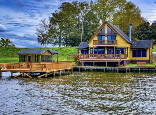 A large yellow house with a deck by the water, alongside a wooden dock and lush greenery in the background.