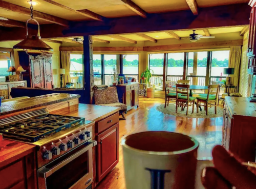A cozy kitchen with wooden beams, a stove, and a view of a sunny outdoor space through large windows.