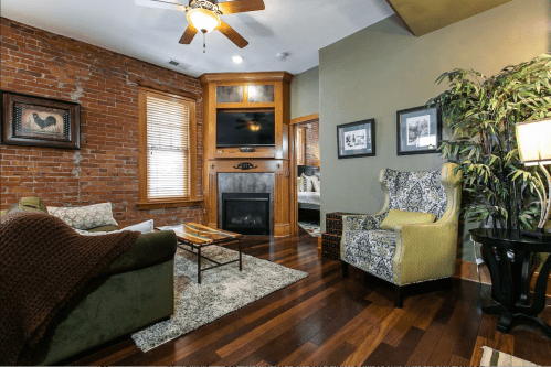Cozy living room with exposed brick walls, a fireplace, and stylish furniture, featuring a green sofa and patterned armchair.
