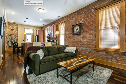 Cozy living room with a green sofa, wooden coffee table, and exposed brick walls, featuring warm lighting and a kitchen area.
