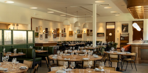 A modern restaurant interior with wooden tables, green seating, and framed artwork on the walls.