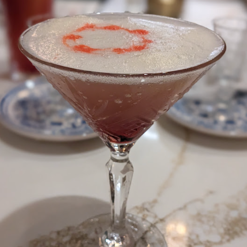 A close-up of a pink cocktail in a crystal glass, topped with frothy foam and a decorative red swirl.