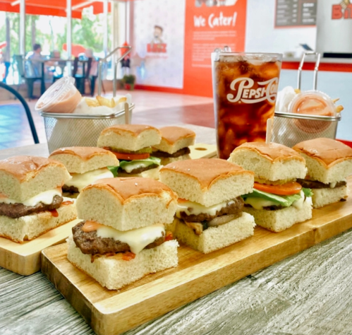 A wooden platter with mini burgers, topped with lettuce, tomato, and cheese, alongside a glass of Pepsi.