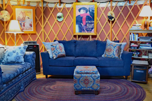 Cozy yurt interior featuring a blue couch with patterned pillows, a small ottoman, and warm lighting.