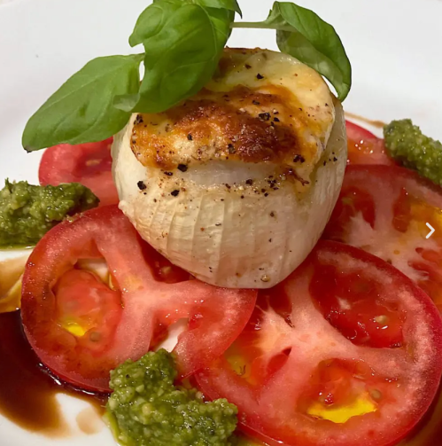 A baked onion topped with cheese, served on sliced tomatoes and drizzled with pesto and balsamic reduction.