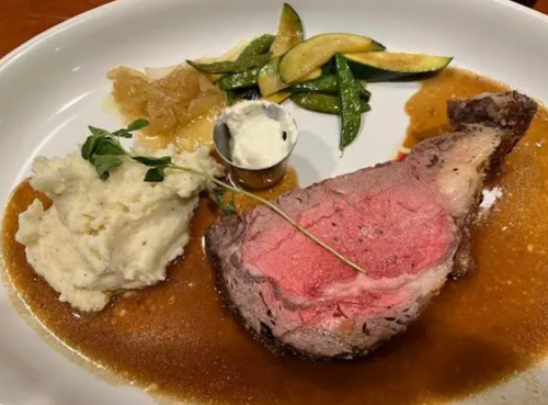 A plate featuring a slice of medium-rare roast beef, mashed potatoes, green beans, and a small dish of sauce.