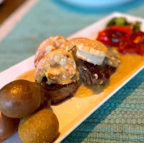 A plate featuring grilled shrimp on steak, accompanied by colorful vegetables and round bread rolls.