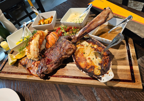 A wooden platter featuring a variety of grilled meats, seafood, and sides, including lobster, shrimp, and baked potatoes.