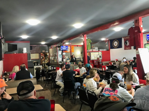 A busy diner with patrons at tables, red accents, and a menu board in the background. Casual dining atmosphere.