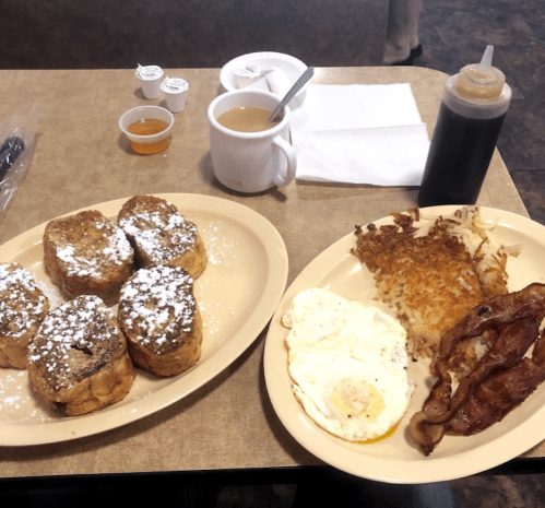 A breakfast spread featuring French toast with powdered sugar, eggs, hash browns, bacon, coffee, and syrup.