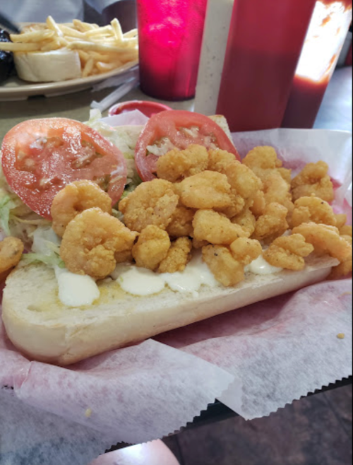 A sandwich filled with fried shrimp, lettuce, and tomato on a hoagie roll, served with fries in the background.