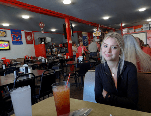 A young woman sits at a table in a casual diner, with drinks in front of her and a bustling background.