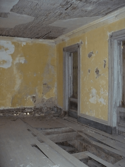 Abandoned room with peeling yellow paint, exposed floorboards, and damaged walls, creating a sense of decay.