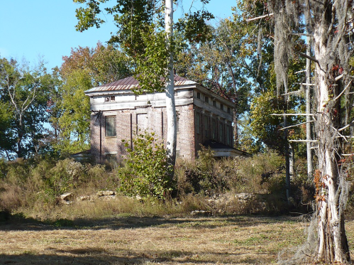 Abandoned brick building surrounded by trees and overgrown vegetation on a sunny day.
