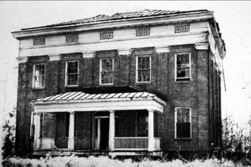 Historic two-story brick house with a decorative frieze, front porch, and a sloped roof, surrounded by overgrown vegetation.