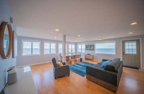 Bright, airy living room with large windows overlooking the ocean, featuring a sofa, chairs, and a dining table.