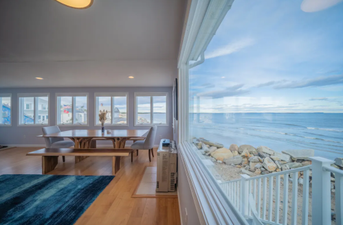 Bright coastal dining area with a wooden table, large windows, and a view of the ocean and rocky shoreline.