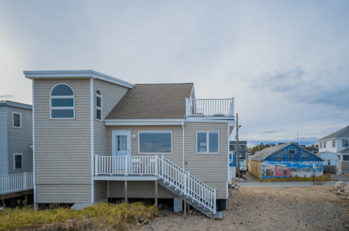 A coastal house with a deck, surrounded by sandy terrain and nearby colorful buildings under a cloudy sky.