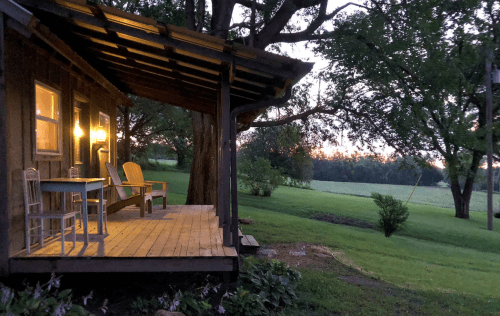 A cozy cabin porch at dusk, with chairs and a table, surrounded by lush greenery and a soft sunset glow.