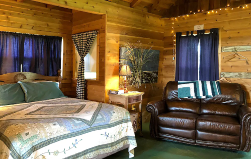 Cozy wooden cabin interior featuring a bed with a quilt, a leather couch, and warm lighting.