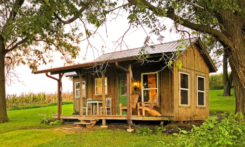 A rustic wooden cabin with a porch, surrounded by greenery and trees, set against a backdrop of cornfields.