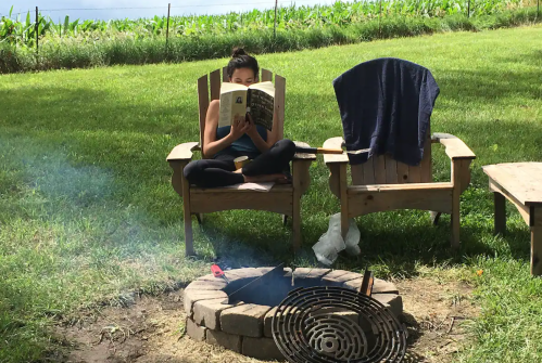 A person sits in an outdoor chair reading a book by a fire pit, with a towel draped over another chair nearby.