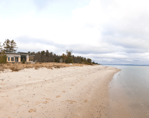 A serene beach scene with soft sand, gentle waves, and a modern house surrounded by trees under a cloudy sky.
