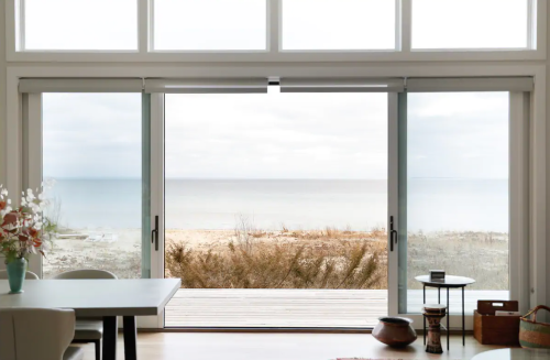 A serene view of a beach through large sliding glass doors, with a dining area in the foreground.
