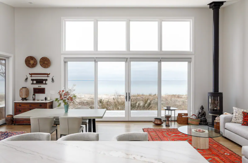 Modern coastal living room with large windows, a dining area, and a view of the beach. Warm decor and natural light.