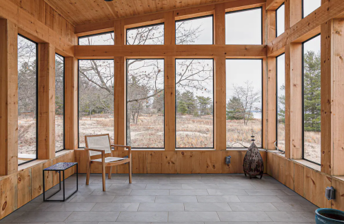 A bright sunroom with large windows, wooden walls, a chair, and a small table, overlooking a natural landscape.