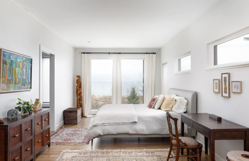 Bright, modern bedroom with a large window, a bed with pillows, a wooden desk, and decorative art on the walls.