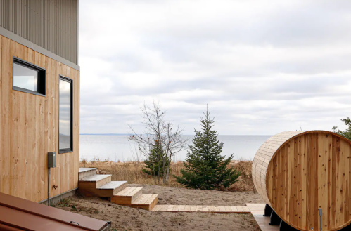 A wooden cabin with a hot tub, steps leading to a sandy area, and a view of a calm lake under a cloudy sky.
