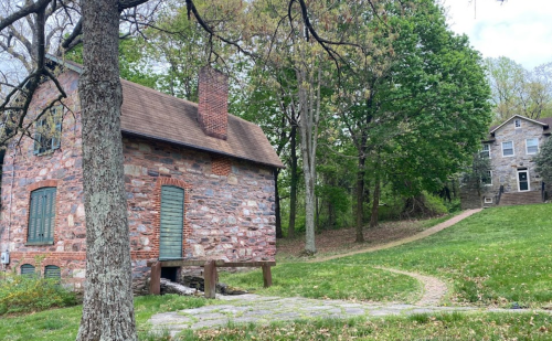A stone building with green shutters beside a grassy path leading to another stone house in a wooded area.
