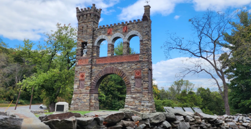 Stone archway with towers and decorative elements, surrounded by trees and a blue sky with clouds.