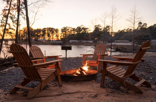 A cozy campfire surrounded by wooden chairs by a lake at sunset, with trees in the background.