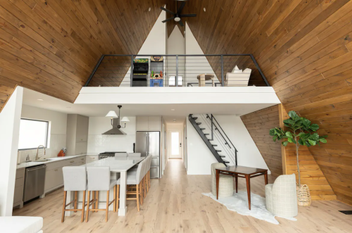 A modern A-frame interior featuring a kitchen, dining area, and loft with wooden walls and a staircase.