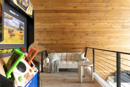 A cozy interior with a wooden wall, a beige couch, and a vintage arcade game in the foreground.