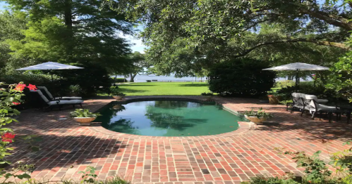 A serene backyard with a pool surrounded by brick, lush greenery, and shaded seating areas under umbrellas.