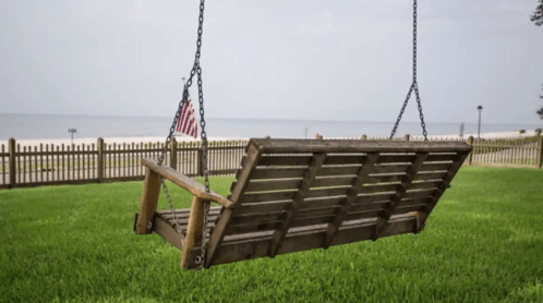 A wooden swing hangs from chains in a grassy area, overlooking a calm beach with a fence and an American flag nearby.